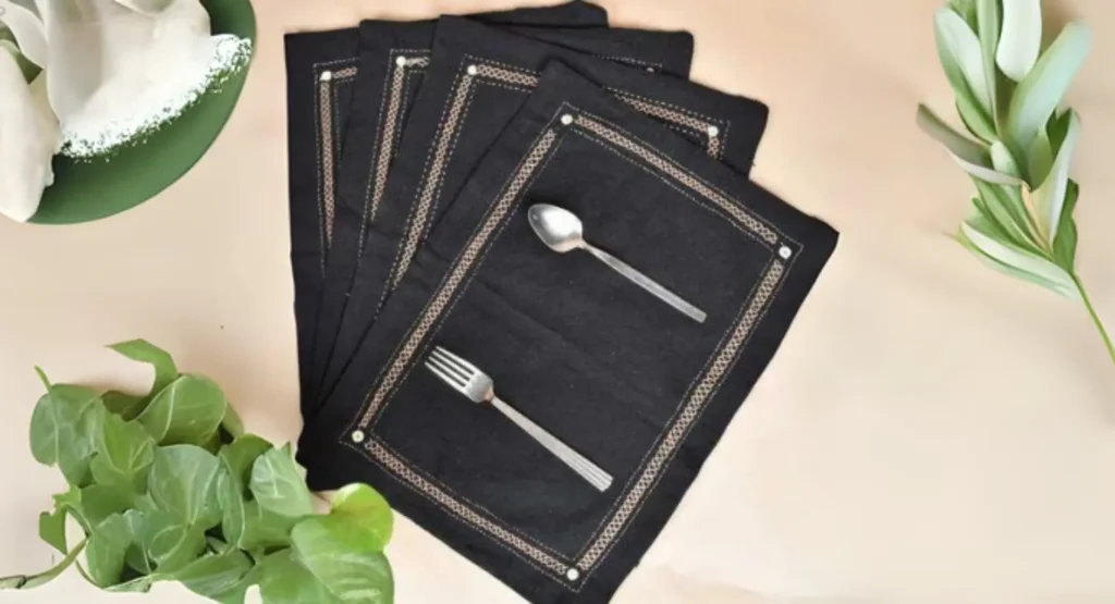 Set of four black fabric placemats with gold trim, modern design, styled with cutlery, greenery, and a white napkin.